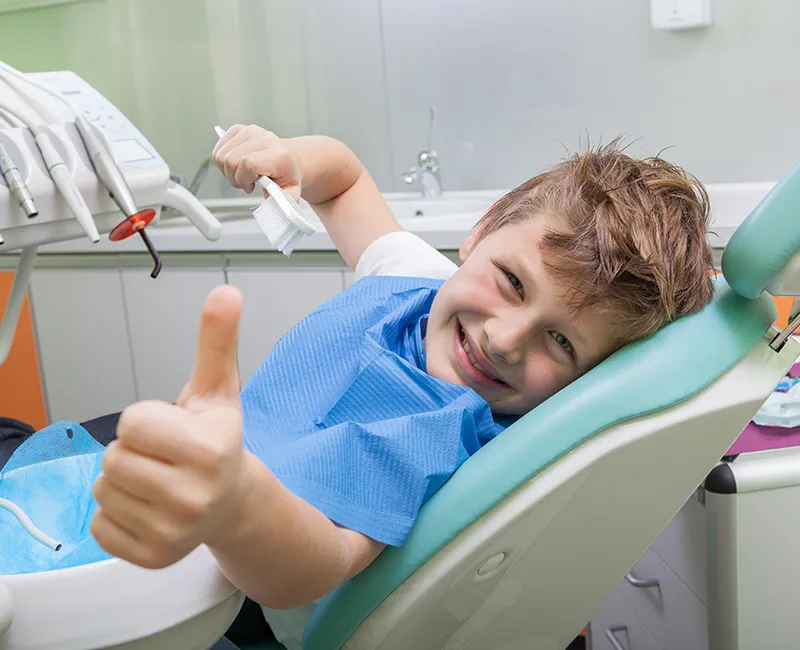 Kleiner Junge beim Zahnarzt zeigt Daumen nach oben während er in die Kamera schaut und auf einem Stuhl sitzt